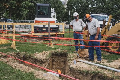 Electrical workers install conduit in a trench while wearing masks.