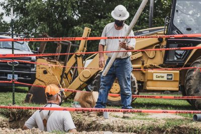 Electrical workers install conduit in a trench while wearing masks.