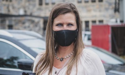 Portrait of someone in a black mask in front of a building and cars. She has blue eyes and long brown hair, wearing a light pink shirt.