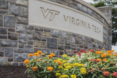 A photo taken down low to the ground showing a bed of orange and yellow flowers in the foreground with a hokie stone sign behind that says "Virginia Tech"