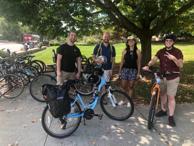 Four people standing with bikes under a tree with a bike rack on the left
