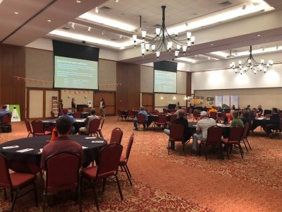 People sitting at large round tables in a conference room with two large screens on the back wall