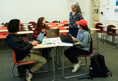 College students sitting at a table in a classroom talking to someone standing