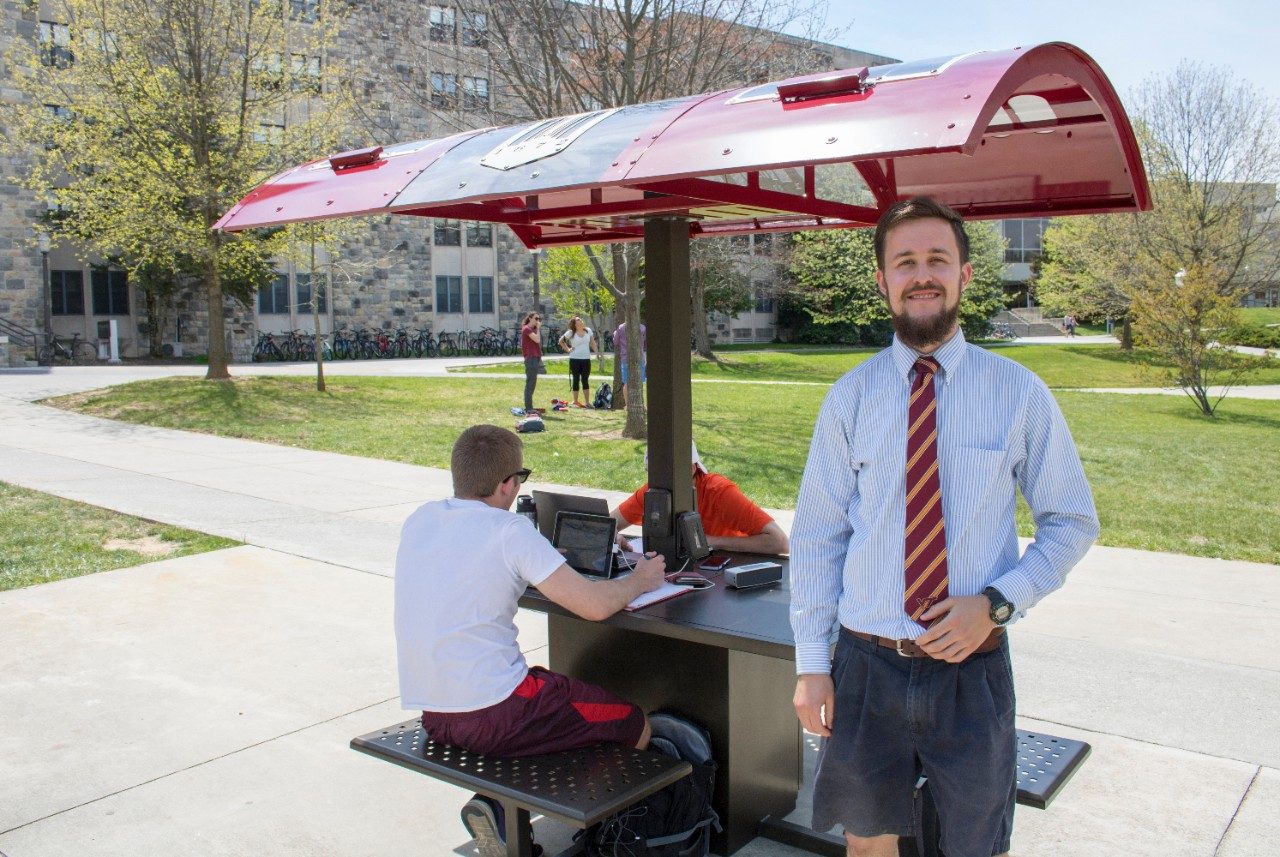 The student inventor of the solar table posing in front of the table with two students using it to charge their laptops as they study.