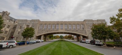 A cloudy blue sky is behind Torgersen Bridge while the grass underneath is bright green. Cars line the street below.