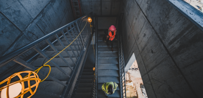 A dark stairway on a construction site with one person wearing bright pink PPE and one wearing orange and yellow PPE.
