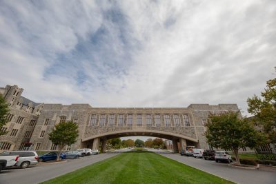 A cloudy blue sky is behind Torgersen Bridge while the grass underneath is bright green. Cars line the street below.