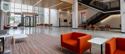 Inside the entrance of Hitt Hall with orange chairs and tables around a large space, with a lit stairway going up in the background.
