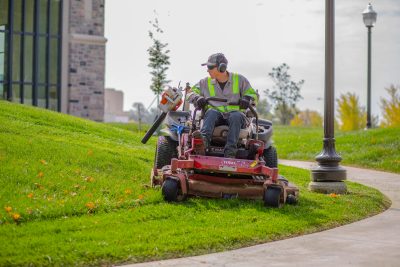 A person wearing high vis clothing, ear protection, gloves, and glasses drives a riding lawnmower with a grey Hokie Stone building in the background
