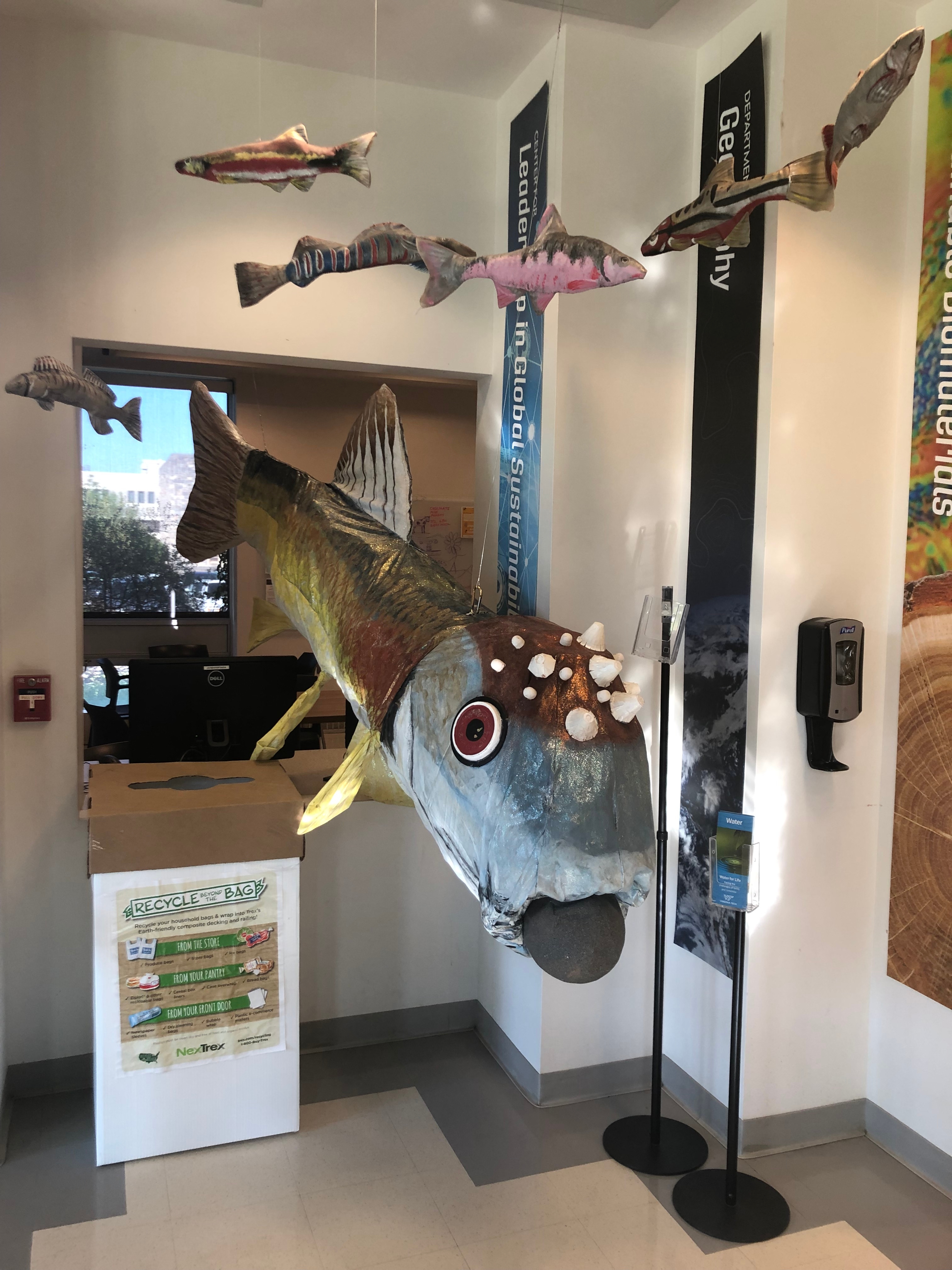 A white soft plastic recycling bin is placed to the left of a large model fish on the first floor of Cheatham Hall.