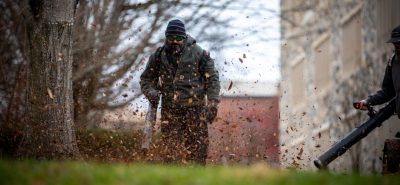 A man wearing safety sunglasses uses a leaf blower to scatter a spray of autumn leaves on the campus grounds.