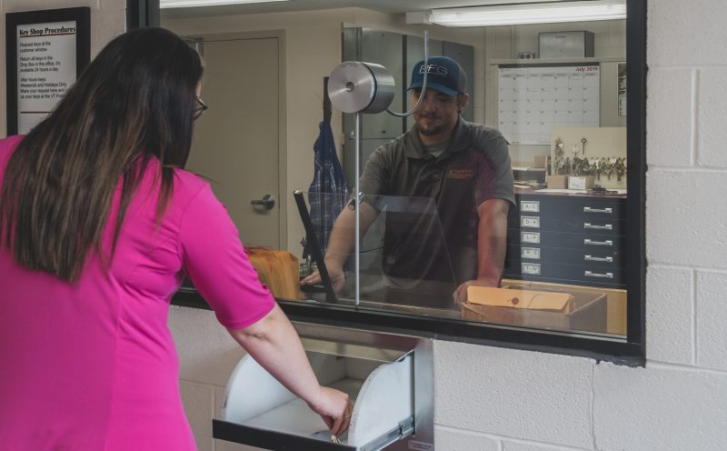 A customer places a key into a slide out drawer at the customer service counter of the Access Control Shop. She is being helped by a young man wearing a hat, standing behind glass inside the shop where the keys are made.