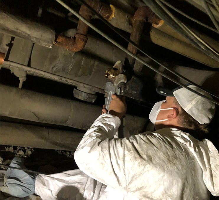 A Facilities worker in a white Tyvek suit, mask, and booties is using a large pipe wrench to turn a pipe connector while seated in a crawl space under Burruss Hall..