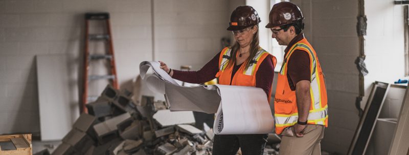 Two people in maroon hard hats and safety vests read construction plans inside a room that has been recently demolished.