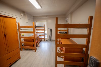 A dorm room with white walls, gray floor, new wooden furniture and a small window in the corner