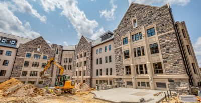 A wide angle view of a campus building under construction, with an excavator digging around in a large area of dirt.