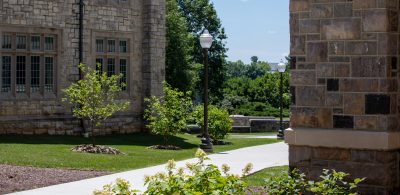 A sunny summer day with green trees and a sidewalk meandering through hokie stone buildings on campus