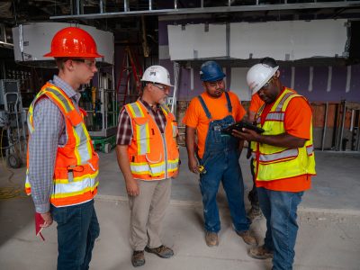 Several people wearing construction PPE gather together on a job site to examine a plan on an iPad