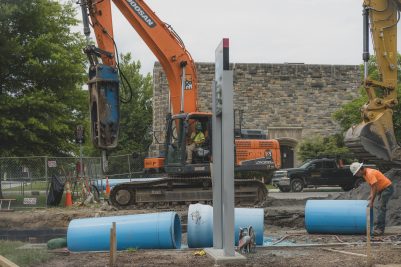 Many large blue pipes alongside a trench being dug by heavy equipment near grey Hokie Stone buildings