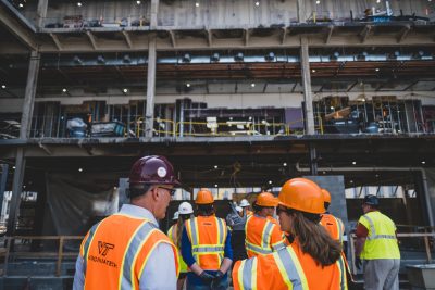 A group of people wearing orange high visibility vests and hard hats stand in front of an active construction site. 