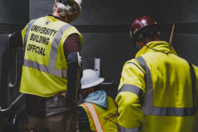 Two people wearing yellow safety vests and hard hats peer over at someone also wearing a yellow safety vest and a hard hat who is measuring a toilet in an ADA bathroom. One of the people looking over has a vest that says University Building Official on the back of it.
