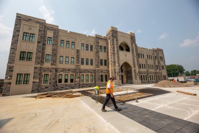 Grey Hokie Stone Corps Leadership and Military Science Building under blue skies. A person wearing a high visibility vest and hard hat walks in front of the building. 