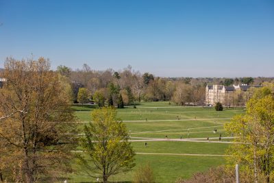 Students walk across the Drillfield on a fall day. A grey Hokie Stone building is in the distance. Trees line the Drillfield. 