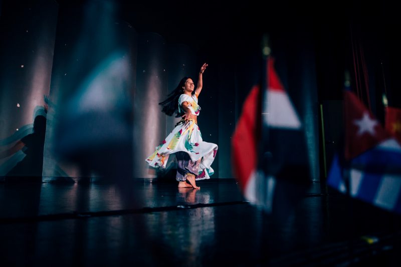 A woman, barefoot, in flowing purple pants, a white and floral skirt, and a matching white and floral top dances with one arm raised and another outstretched towards the camera. She is in front of a brightly lit blue-grey backdrop with out-of-focus flags of multiple nations in the foreground.