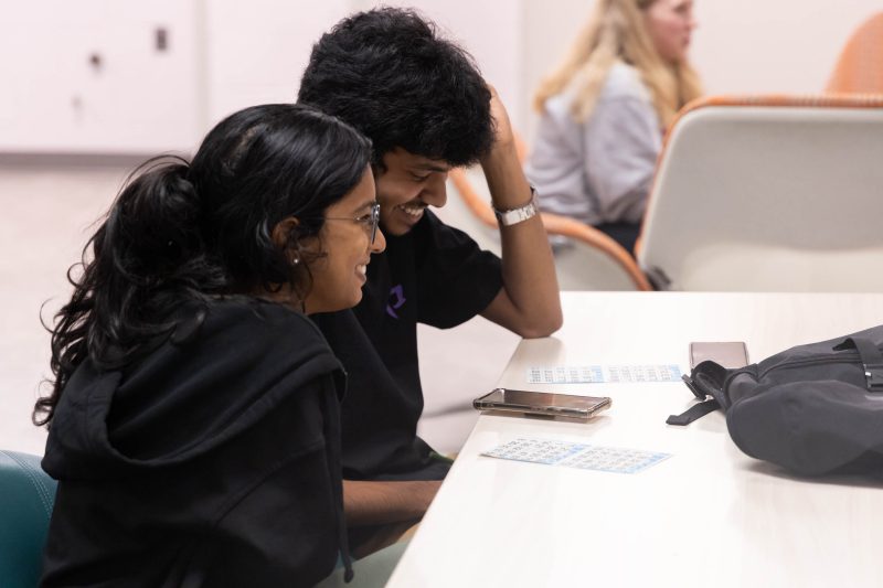 Two students in casual dress sit at a grey table, smiling, with a cell phone, a backpack, and partially marked Bingo sheets on the table in front of them.