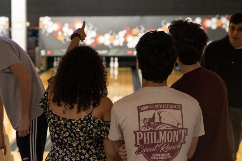 Three students stand with their backs to the camera, pointing offscreen. In the background is several lanes of a bowling alley with some pins knocked over and a hexagon pattern on the wall above it.