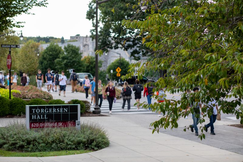 Many young people cross sidewalks at an intersection. In the foreground is a large green tree and a sign that says Torgersen Hall. In the background are grey Hokie Stone buildings and more green trees.