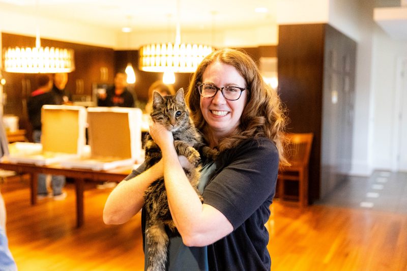 Ashley Reed smiles as she holds her cat Ava in West Ambler-Johnston hall with students enjoying pizza in the background.