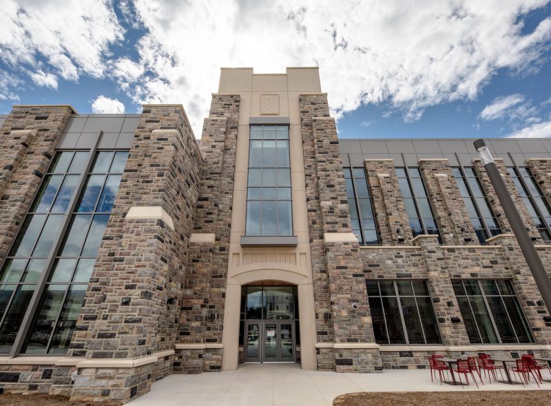Front view of Hitt Hall constructed with Hokie Stone and large vertical windows. The entrance features a tall central section with glass doors beneath a stone archway. The sky above is partly cloudy with patches of blue. To the right, red chairs and tables are arranged on a concrete patio area