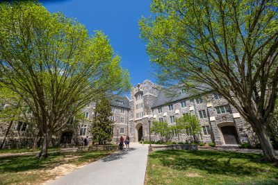 Students walk along a paved pathway toward grey Hokie Stone Eggleston Hall with a central arched entrance. The building is framed by leafy green trees on both sides under a clear blue sky, creating a bright and welcoming spring atmosphere.