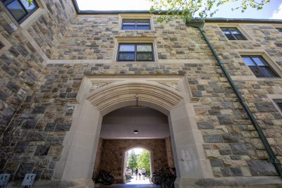 A view looking up at grey Hokie Stone Vawter Hall, featuring a wide arched passageway with decorative stonework above the entrance. Several bicycles are parked inside the archway, and three people are walking through it toward a sunlit green space beyond.