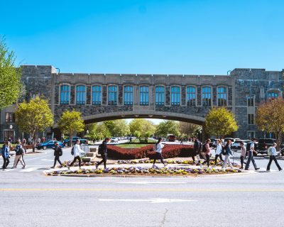 Students walk across a crosswalk lined with colorful flowerbeds and a bush shaped like the Virginia Tech logo on a sunny day at Virginia Tech, with grey Hokie Stone Torgersen Hall bridge and its large arched passageway in the background. 