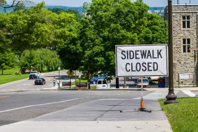 Signage reading "Sidewalk Closed" is placed on a sidewalk on campus. Grey Hokie Stone Agnew Hall is in the distance. Cars travel on the tree lined road adjacent to the sidewalk.