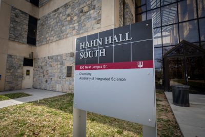 Black, grey, and maroon sign for Hahn Hall South. Chemistry and Academy of Integrated Science are listed on the sign. Surrounding the sign is a grey Hokie Stone building whose focus is blurred out. 