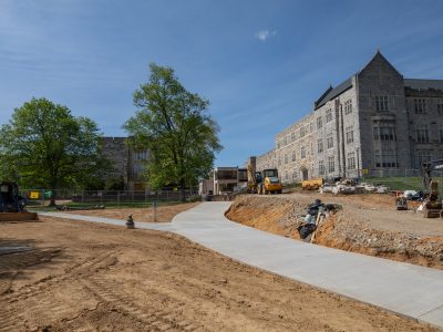 A newly poured concrete sidewalk curves through an active construction site, surrounded by exposed dirt, construction equipment, and fencing. Hokie Stone Pamplin and Burruss Halls, partially obscured by trees, are visible in the background under a clear blue sky.
