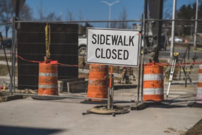Signage reading "Sidewalk closed" in front of a gate and orange traffic cones. 