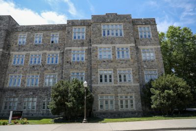A large stone academic building with four stories and rows of white-framed windows stands under a bright blue sky with wispy clouds. The building is surrounded by neatly trimmed shrubs and a well-maintained lawn. A campus sign in the lower left corner reads “Patton Hall, 750 Drillfield Drive.” A lamppost is positioned near the center of the building’s facade.