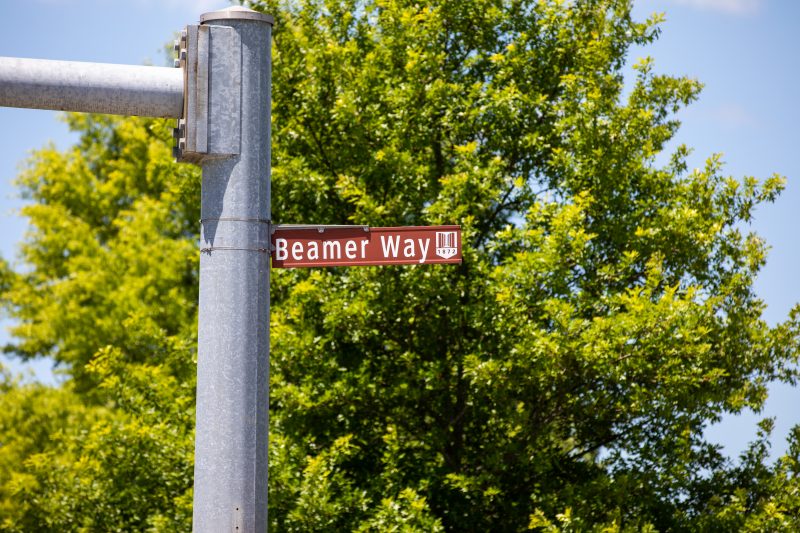 Brown street sign with the Pylons Virginia Tech logo on the end of it that reads Beamer Way. The sign is surrounded by green trees and blue sky.