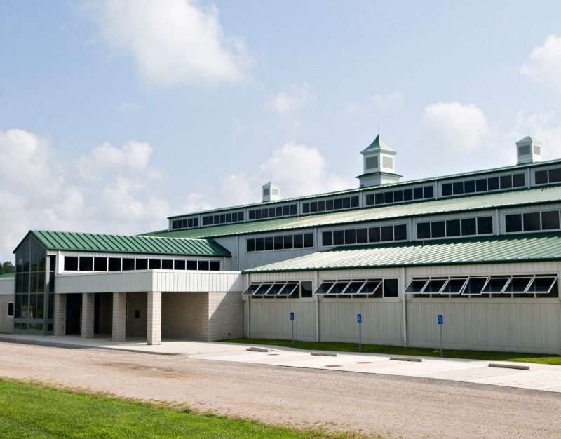 A large white barn with a light green roof under blue skies. A dirt and gravel covered road is in front of the barn. 