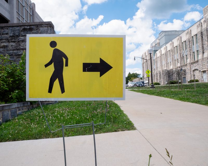 Two young people walk together on a concrete sidewalk. On one side of the sidewalk is grass and lampposts. On the other is a construction fence and construction behind it.