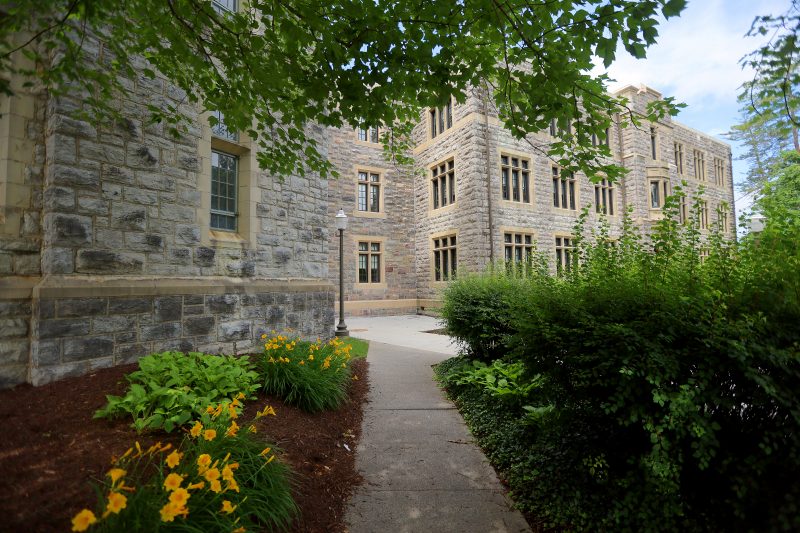 A paved pathway leads up to a grey Hokie Stone building. Yellow flowers and green bushes surround the pathway. 